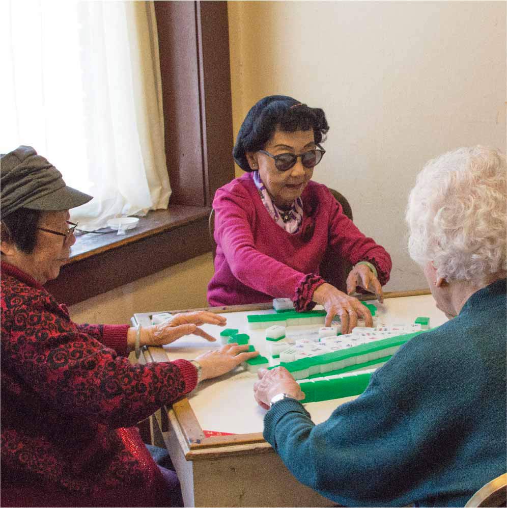 three chinese seniors around a table playing Mahjong. The women in the middle has black hair done in a 50s like manner and is wearing Pantos shaped sunglasses, a pink sweater and pink ascot. She has her hands out on the table to grab a tile.