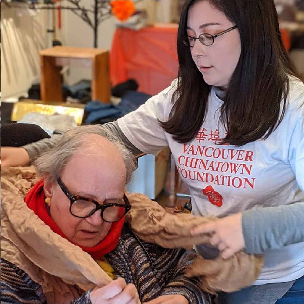 A young woman helping an older woman try on a scarf. The younger woman is on the right side of the photograph and is wearing a Vancouver Chinatown Foundation volunteer t-shirt, glasses, and a long sleeve shirt. The older woman is on the left and is wearing glasses and a large puffy jacket. The volunteer is helping her put on an orange scarf